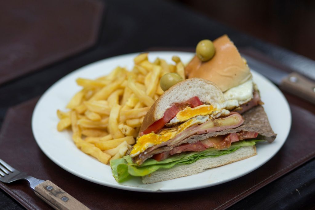 A chivito (Uruguayan sandwich) served with fries on a white plate on the table.